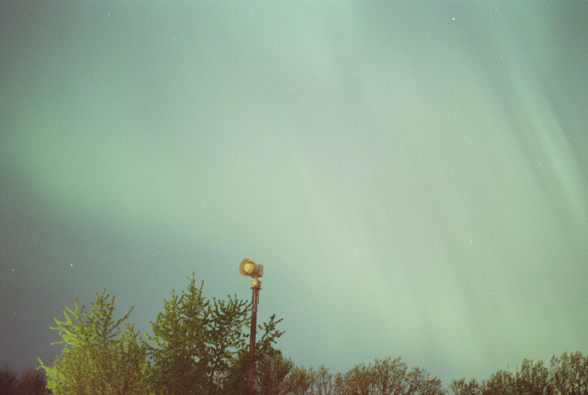 The aurora borealis is visible in the sky above some tree cover. A speaker mounted to a pole is visible in the foreground.